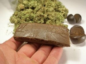 Schon gewusst... HaschischEin Bild, das in Vordergrund auf einer Hand ein großes quadratisches Stück Haschisch, in Hintergrund auf ein Tisch Cannabis Knospen und rechts auf den Tisch und Bild runde in der Mitte durchgeschnittene Haschisch Kugel zeiget.